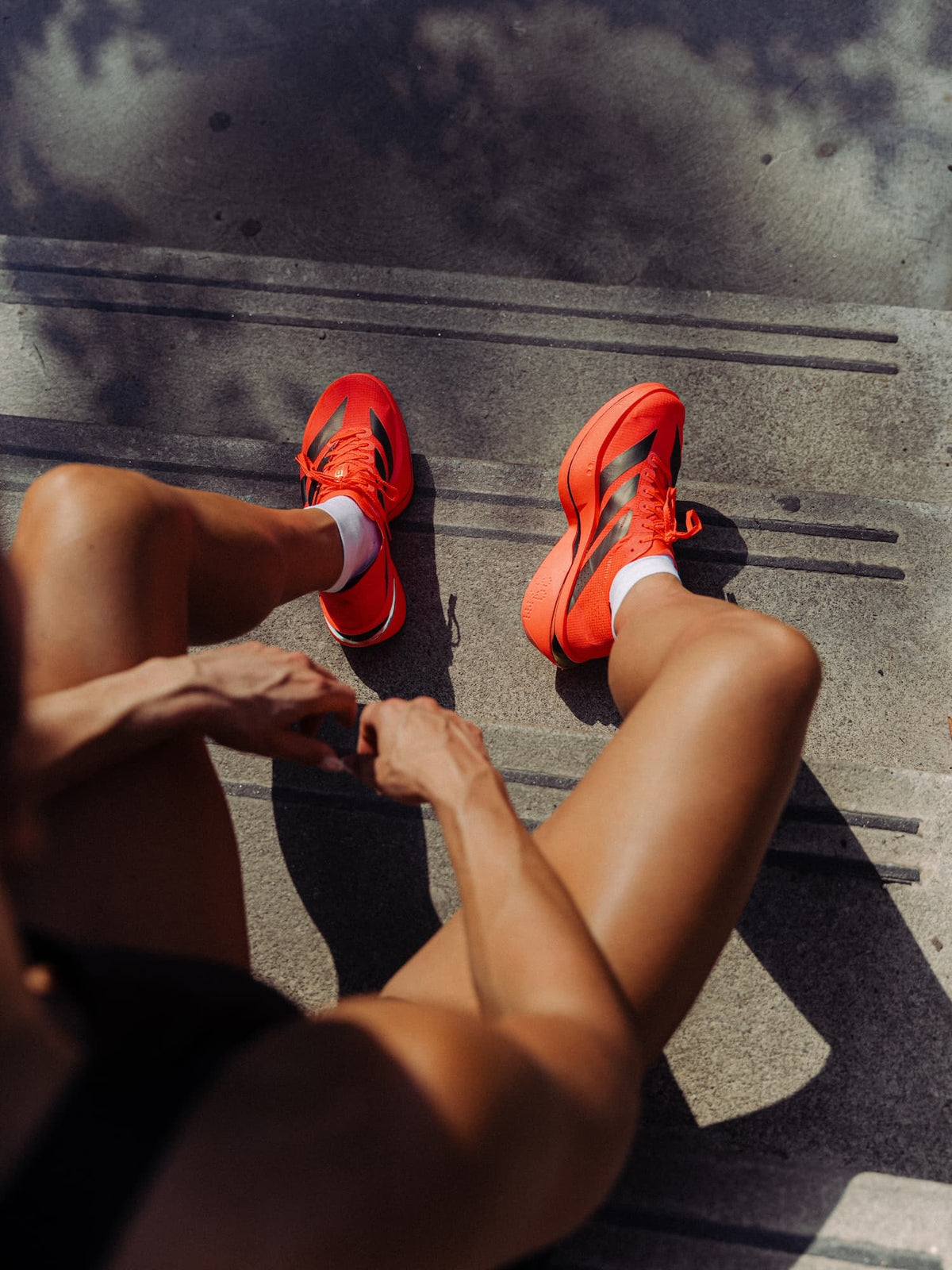 A top-down view of a person sitting outdoors on concrete, wearing bright orange-red Adidas Adios Pro 5 shoes with white socks.
