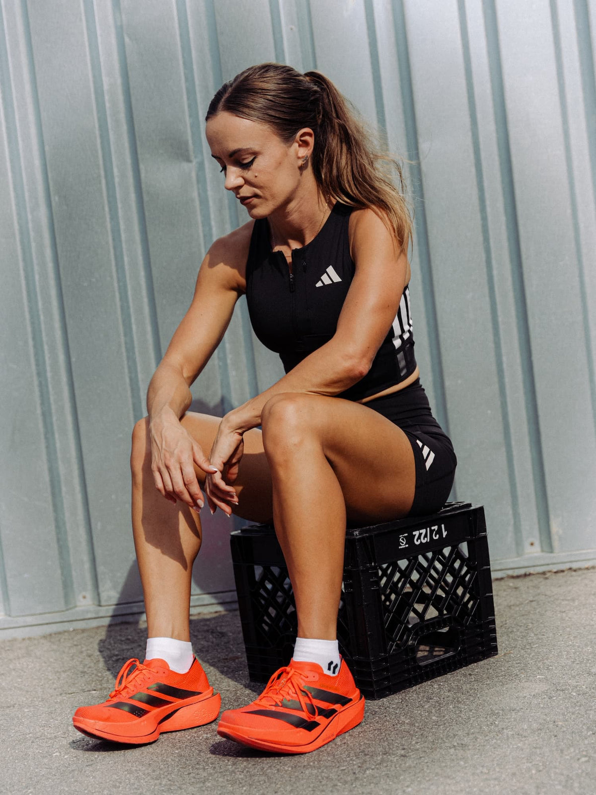 A woman sits on a black plastic crate outdoors against a corrugated metal wall, wearing a black Adidas athletic crop top and shorts with bright orange Adidas Adios Pro 5 running shoes.