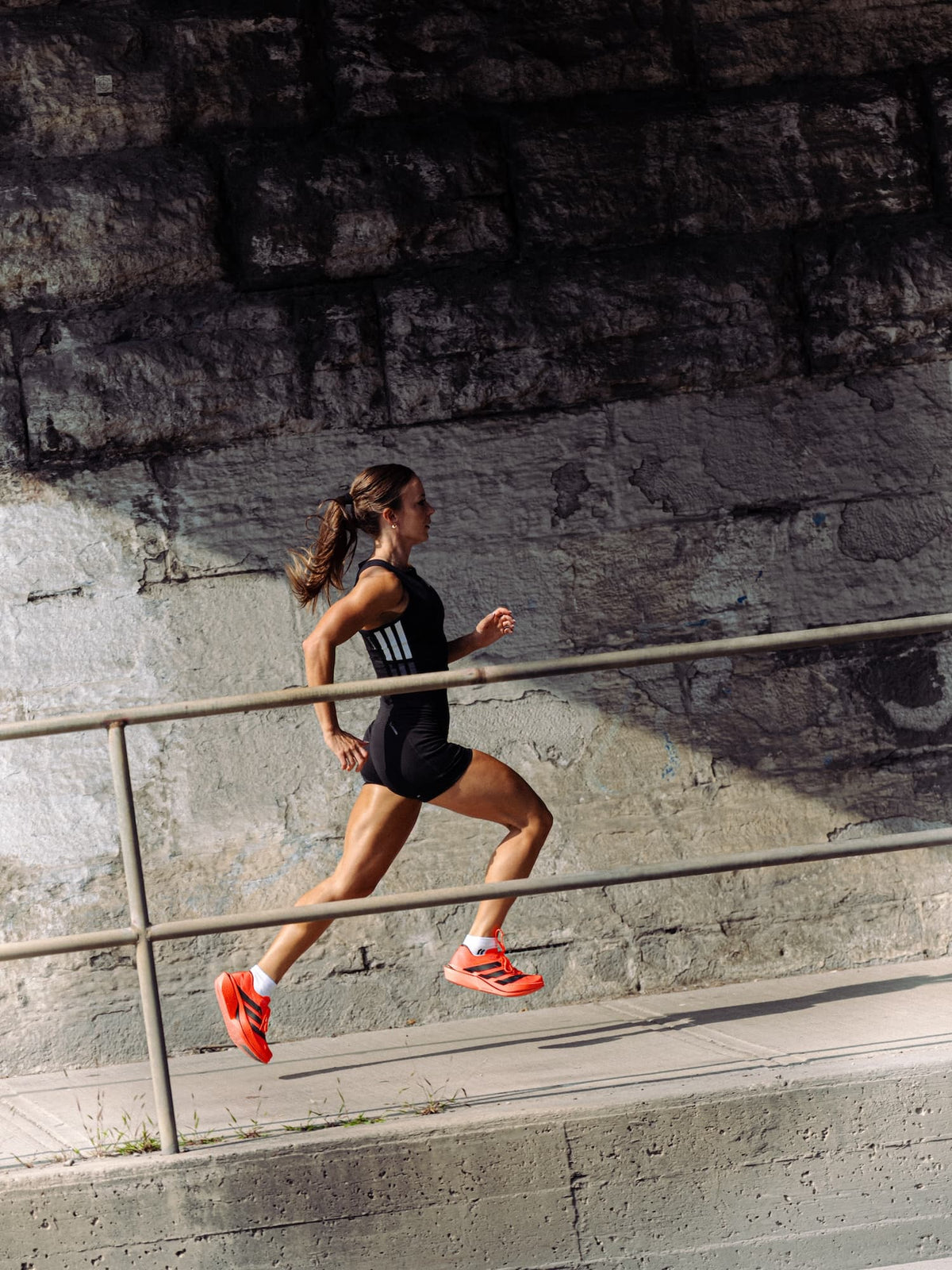 A woman running on a concrete path beside a stone wall, wearing a black sleeveless athletic top with white Adidas stripes, black shorts, and bright orange Adidas adios pro 5 running shoes.