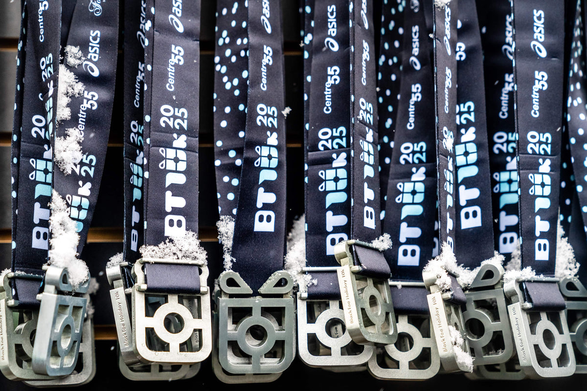 A close-up row of dark blue BT10k lanyards with silver metal medals, each printed with white and blue text including 'BT25', 'Competition 2025', and the Asics logo, with some snow-like dust on the straps.