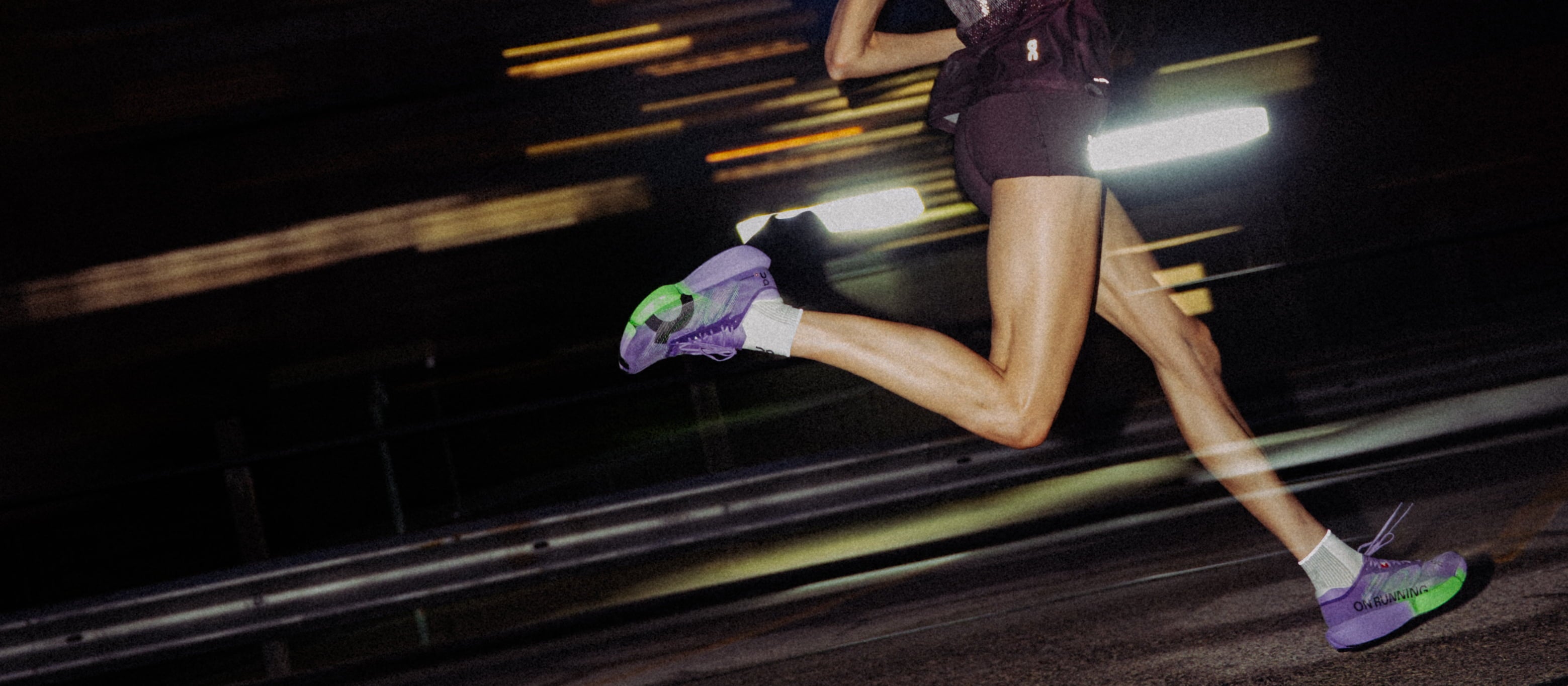 A runner's legs in motion at night, wearing purple and green On Cloudboom Strike 2 running shoes, white socks, and dark shorts, with bright motion blur and streetlights in the background.