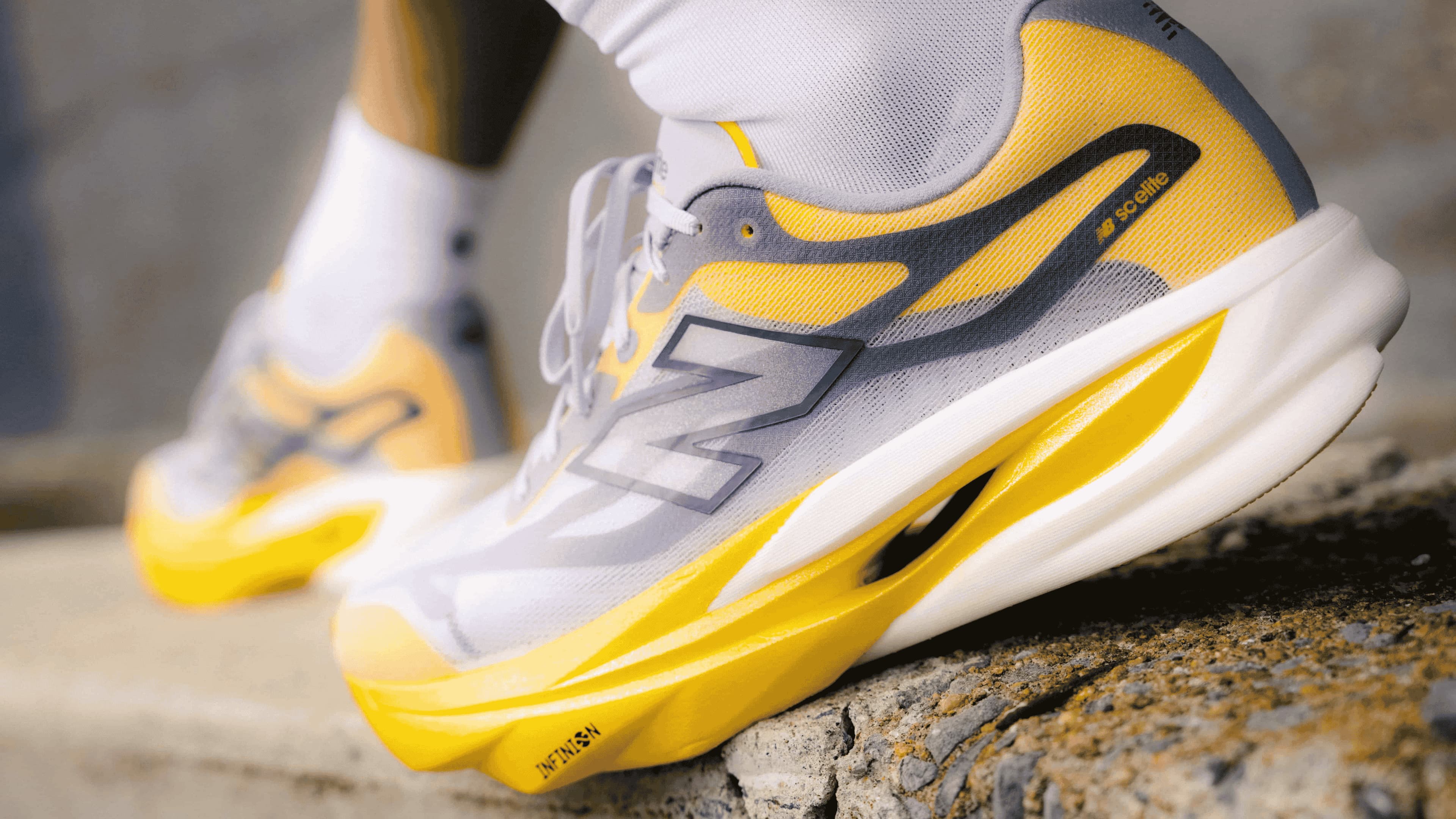 A close-up of a New Balance running shoe being worn, showing the side profile with its signature 'N' logo, yellow and white colorway, and thick cushioned midsole, set against a blurred outdoor background.