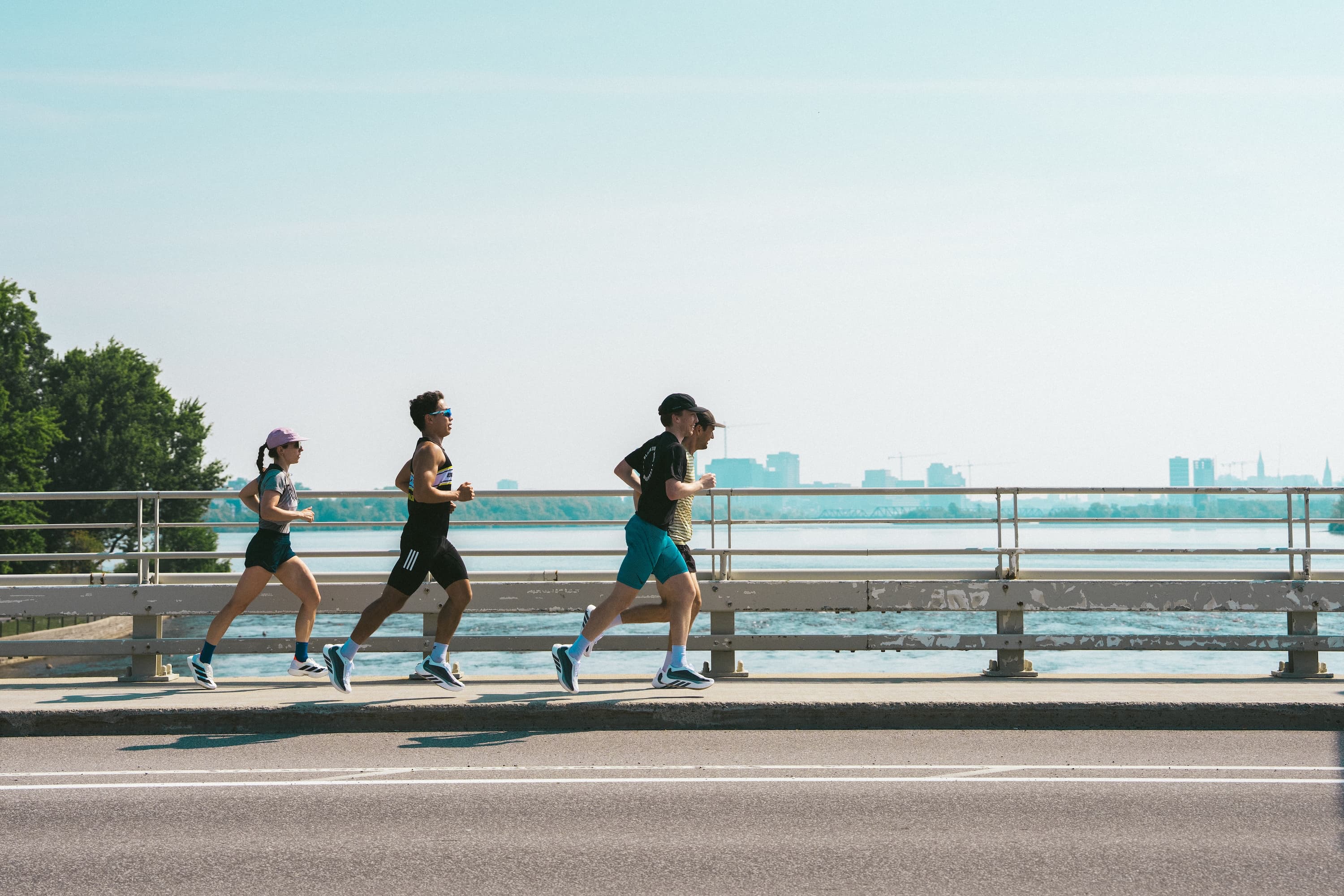 Four runners jogging side by side on a paved road next to a railing with water and a distant Ottawa skyline under a clear blue sky.